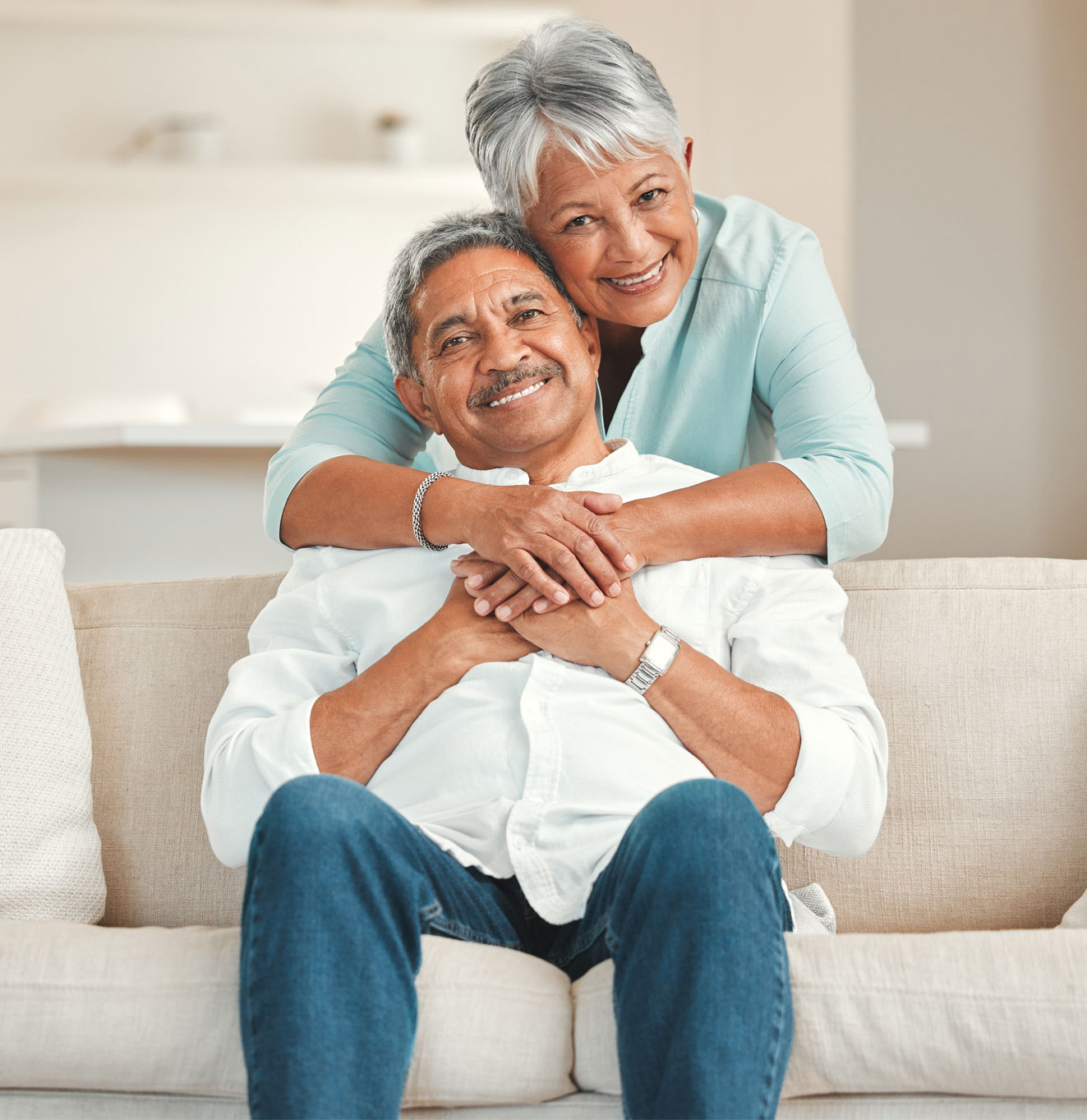 Hispanic couple hugging, the man is sitting on a couch while the woman stands behind him 